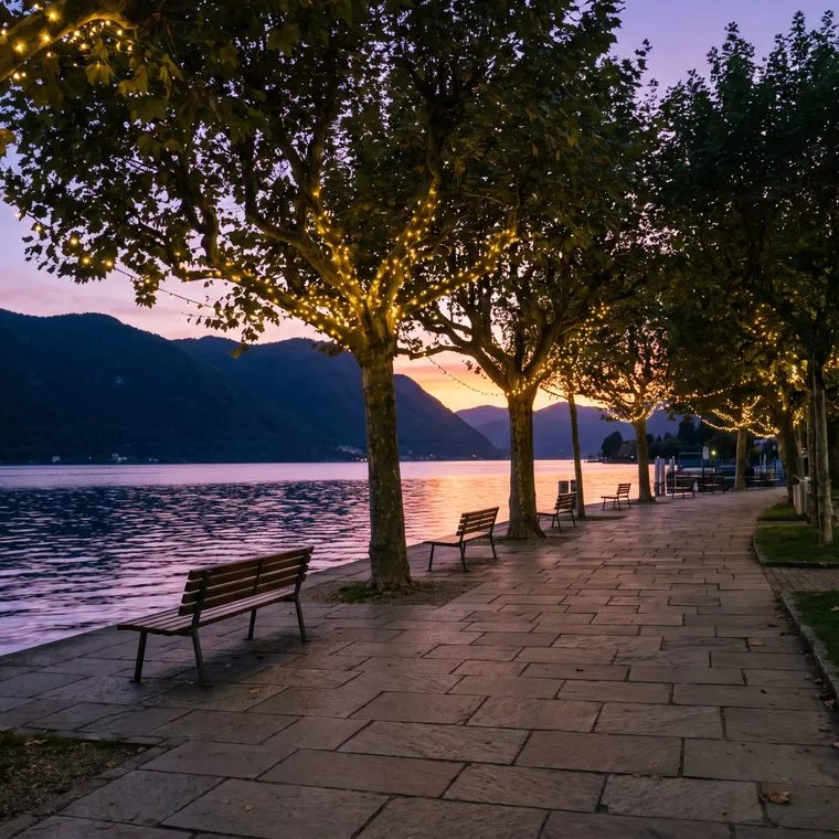 Generated scene: a lakeside promenade at dusk with benches and trees strung with small lights, the lake glowing violet and orange below dark hills.